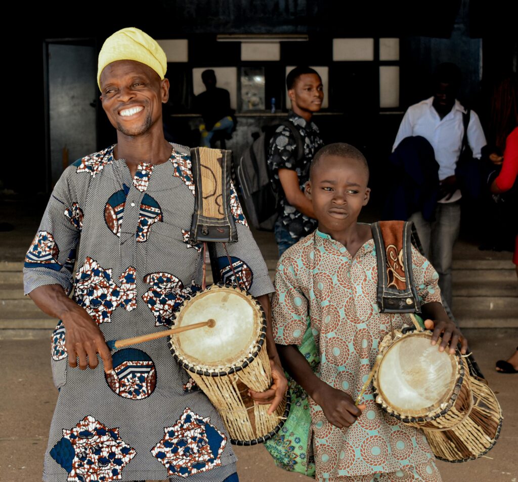 Smiling African drummers in traditional attire playing talking drums outdoors.