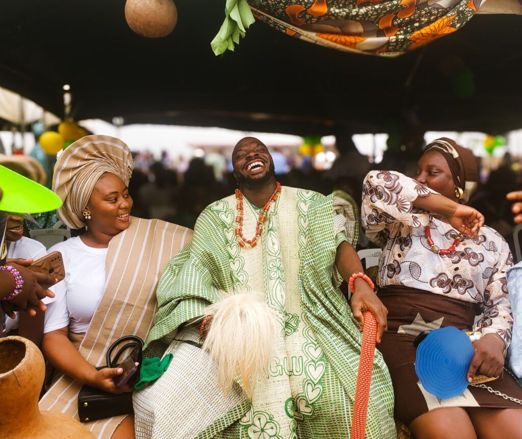 A man in a green patterned agbada laughs out loud, surrounded by smiling friends at a celebration.