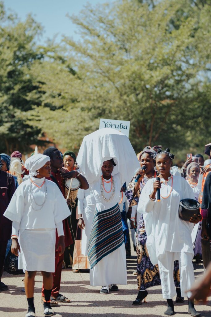 Women in white lead a Yoruba procession; one carries a covered load on her head beneath a sign that reads Yoruba.