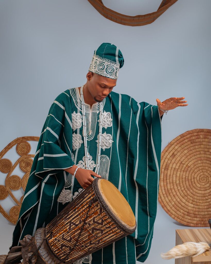A man in a green agbada and beaded cap rests a hand on a carved talking drum, set against woven wall hangings.